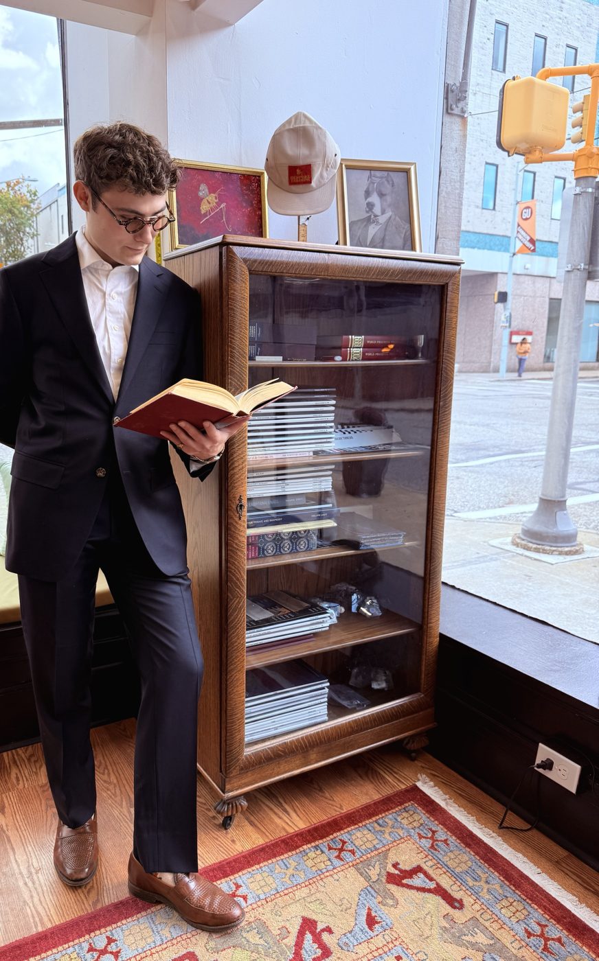 A man in a navy suit reading a book beside an antique glass-front cabinet