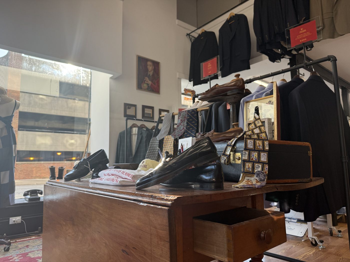 A wooden table with leather shoes, ties and shop t-shirts beside racks of jackets